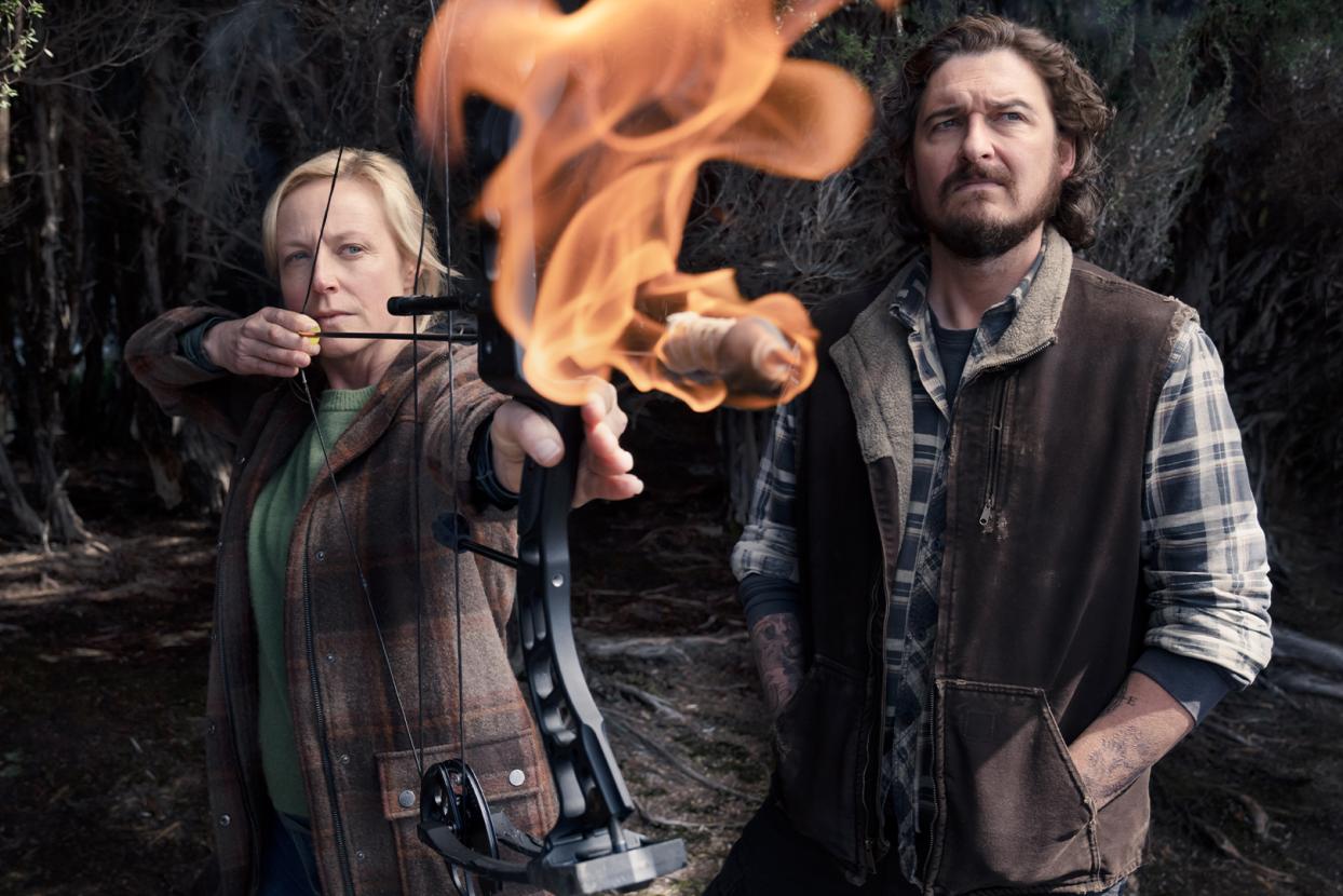 An assured blonde woman (actor Marta Dusseldorp as Stella Heikkinen) stands with a flaming crossbow pulled tautly, beside her a laconic, rural man (acor Toby Leonard Moore, as Jeremiah) wearing a plaid long sleeve shirt and brown coat for warmth, looks out to the distance.